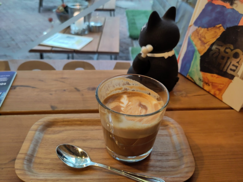 Foto: An einem Fenster eine Holzablage in Tischhöhe, auf der ein rechteckiges Tablet mit eine Flat White in einem Glas sowie ein Kaffeelöffel abgelegt sind. Im Hintergrund eine schwarze Winkekatze sowie ein Buch. Durch das Fenster sieht man einen Tisch mit Sitzgelegenheiten im Freien.