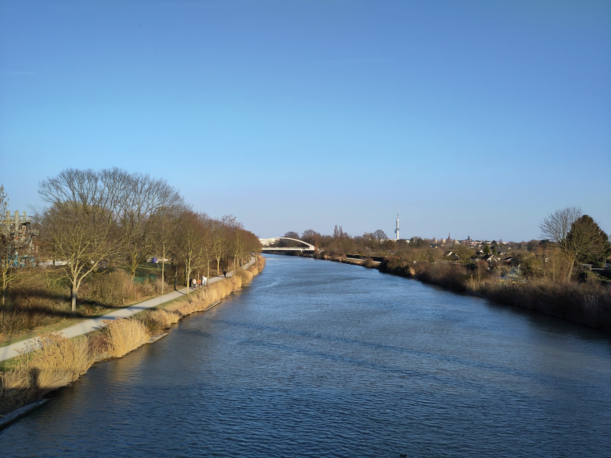 Blick von der Brücke an der Tannenbergallee in Richtung Lister Damm Brücke. Das Wasser im Kanal bildet leichte Wellen, die braunen Bäume und Büsche werden auf der linken Seite von der Sonne beschienen. 