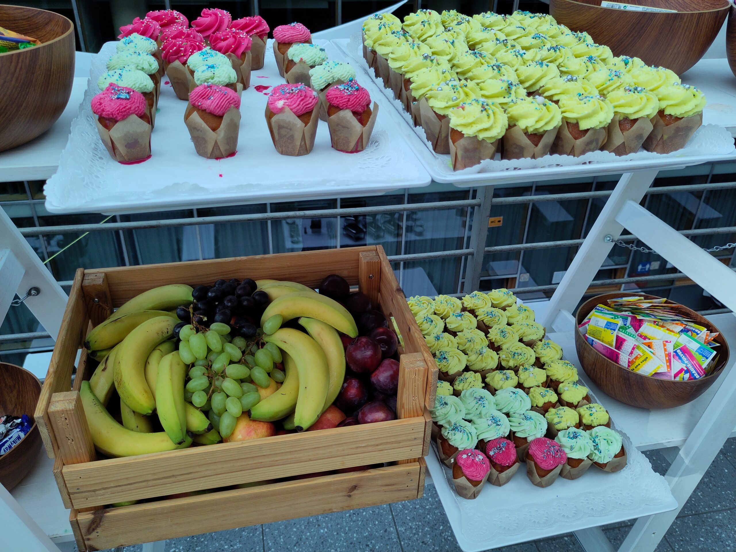 Auf einem zweistöckigen Regal sind Muffins mit unterschiedlicher Creme sowie eine Obstkiste mit Früchten angeordnet.