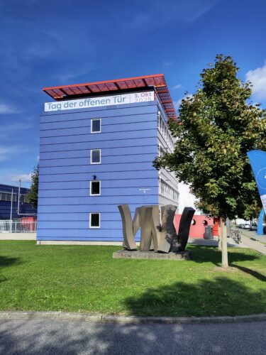 Ein fünfstöckiges Gebäude des Bundesamt für Seeschifffahrt und Hydrographie. Vor dem Gebäude die "tanzenden Pommes", eine Skulptur sowie grüne Bäume.