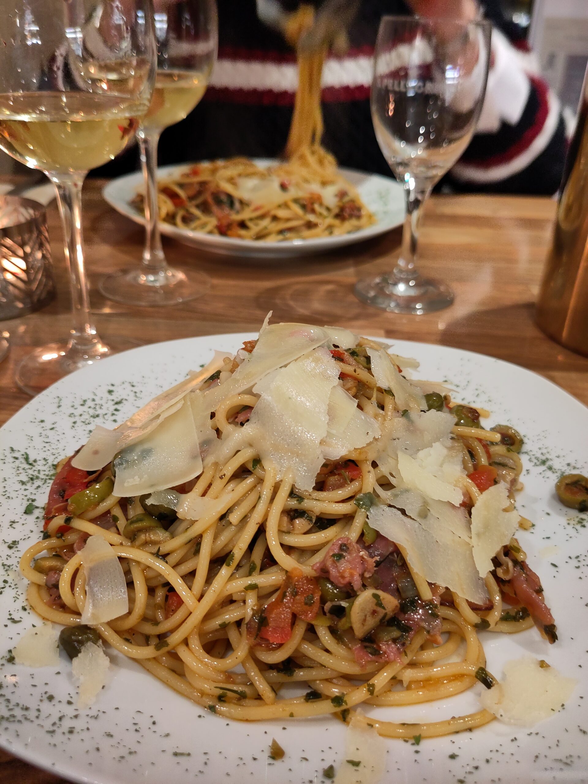 Auf einem Tisch neben Gläsern ein runder Teller mit Spaghetti, die mit Tomatenstückchen, Kapern, Schinkenstreifen und diversen Kräutern vermengt sind. Darauf gehobelter Parmesan.