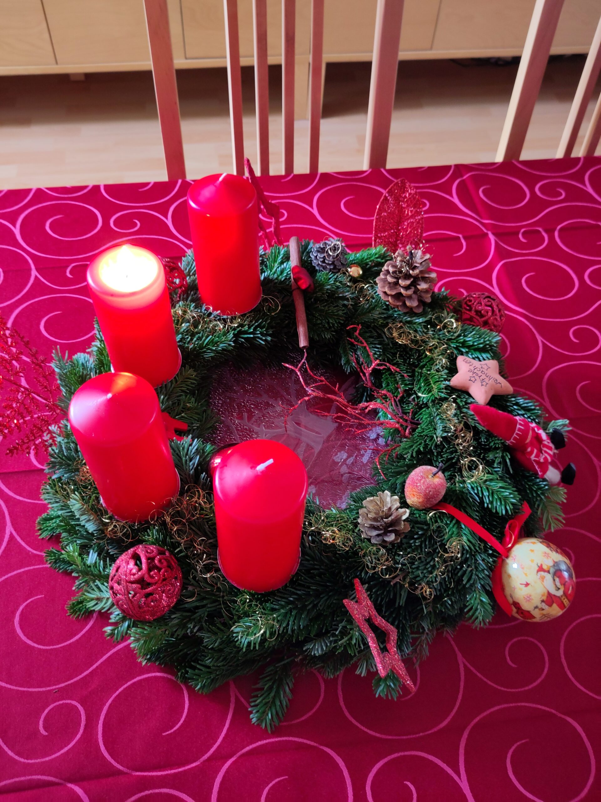 Auf einem Tisch ein runder geschmückter Adventskranz mit vier roten Kerzen. Eine Kerze brennt.