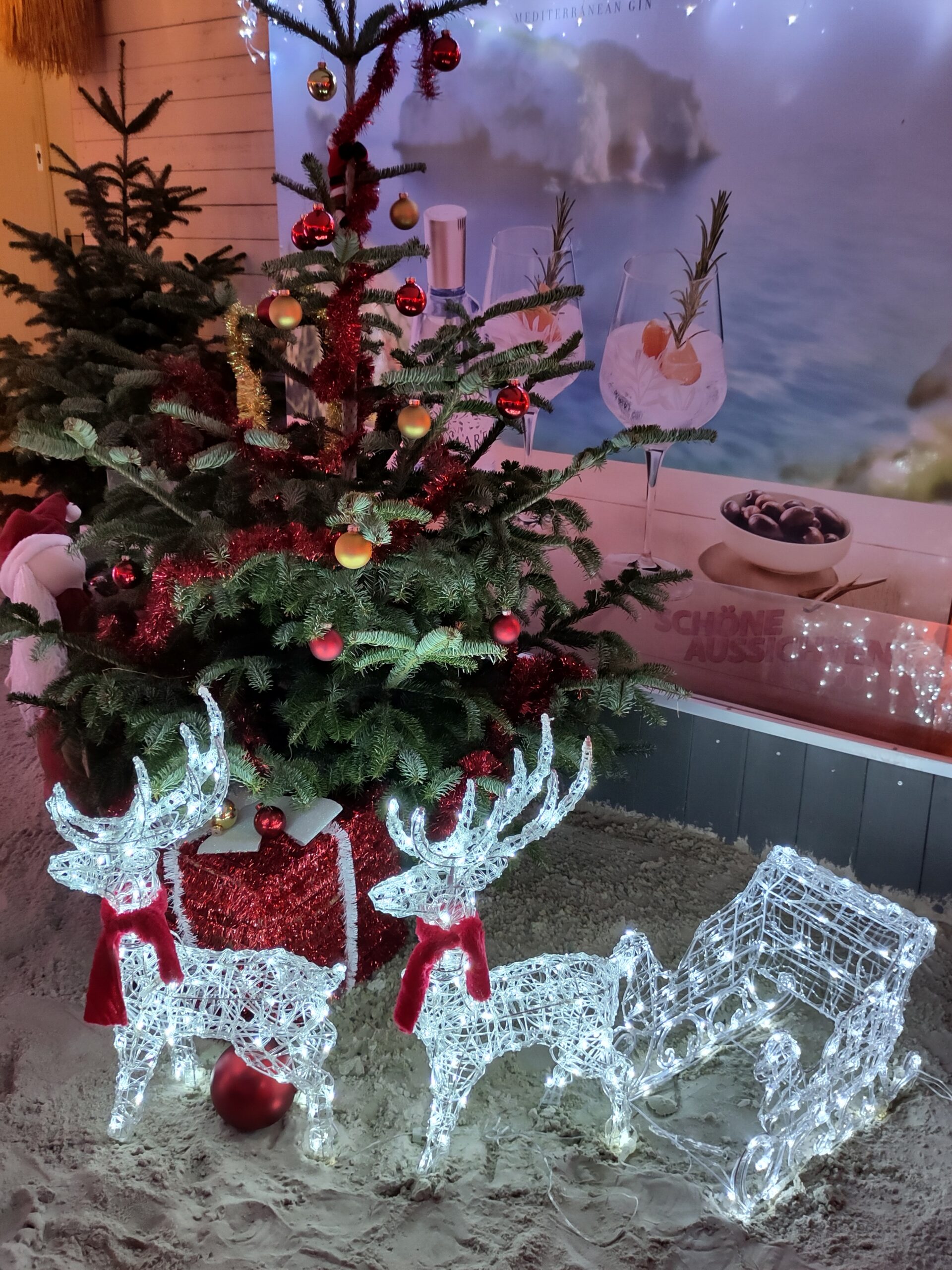 Zwei weiße hell erleuchtete Renntiere ziehen einen weißen hell erleuchteten Schlitten. Daneben ein kleiner geschmückter Tannenbaum. Im Hintergrund an einer Wand ein Plakat von Sommer-Cocktails in Gläsern.