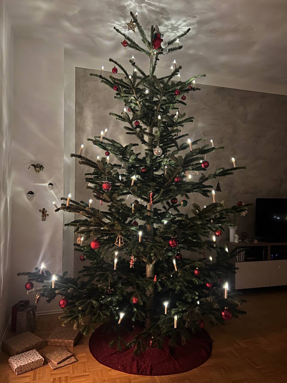 Geschmückter Tannenbaum in der Ecke eines Raumes. Die LED-Lichter am Baum wirken wir Kerzen, die leuchten. Der Ständer des Baums ist mit einer roten runden Decke verdeckt. Links neben dem Tannenbaum auf dem Boden rechteckige, in Geschenkpapiert eingewickelte Gegenstände.