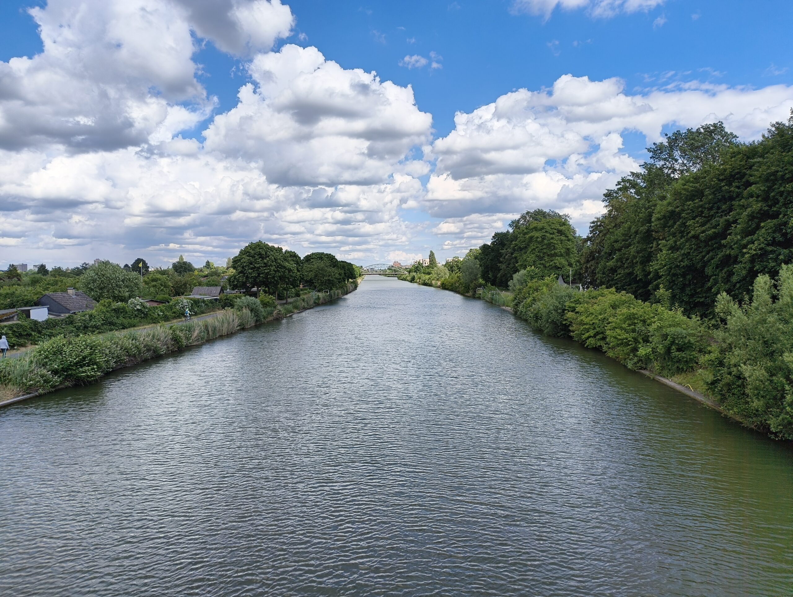Foto: Blick von der Brücke an der Tannenbergallee auf den Mittellandkanal. Das Wasser ist leicht durch gekräuselt, der Himmel blau mit einigen Wolken. Rechts und links sattes Grün durch Wiesenflächen, Büsche, Sträucher und Bäume.