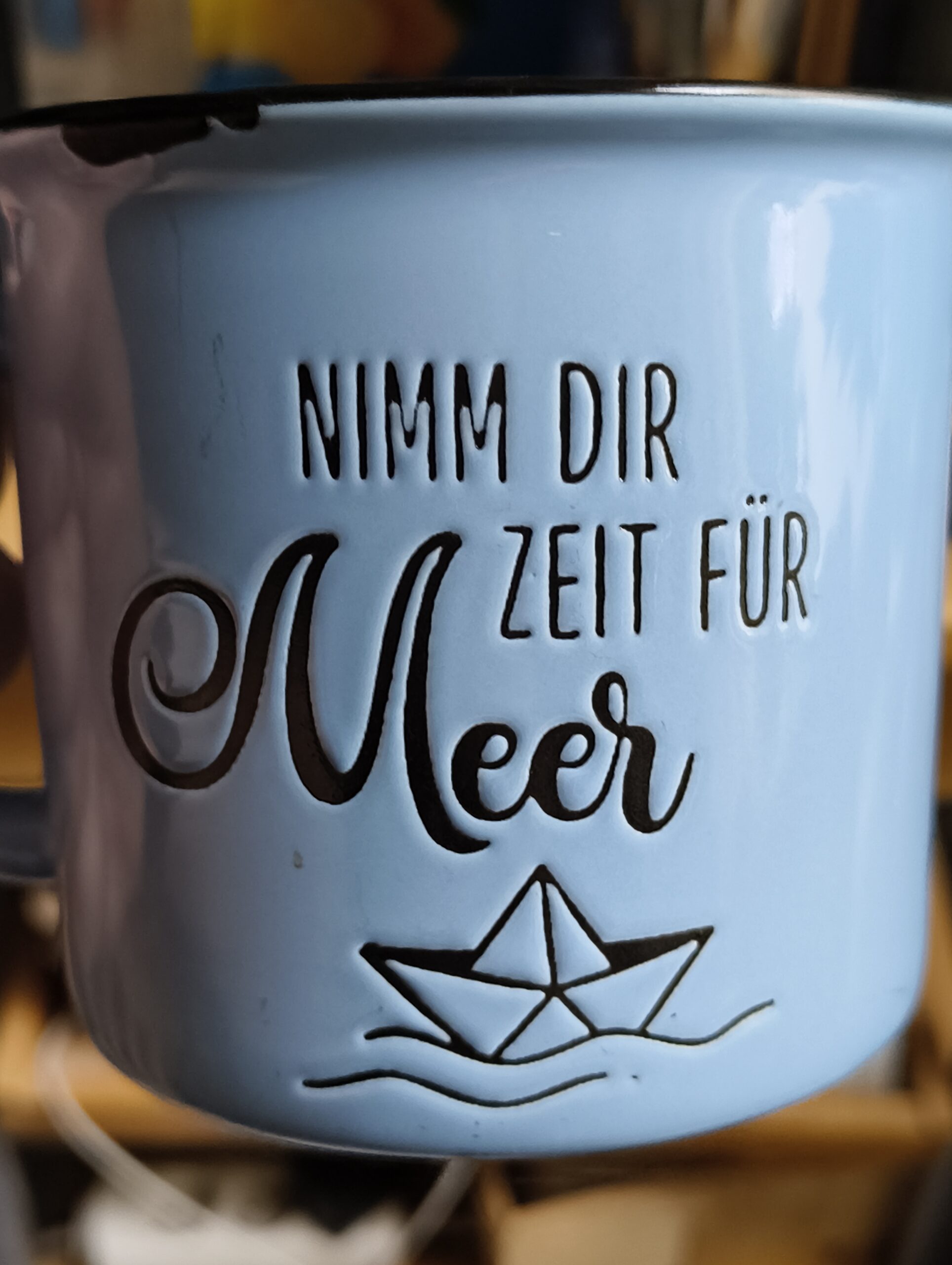 Eine blaue Tasse mit dem Text »Nimm Dir Zeit für Meer«. Darunter ein Papierschiffchen auf Wellen.