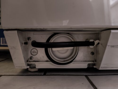 Blick auf die hinter einer klappe verdeckten Zugänge zu einer Waschmaschine. Man sieht ein Einkerbung in einem kreisrunden Element, welches die Verriegelung lösen kann. Man sieht eine größere runde Öffnung mit Steg, über welches man zum Flusensieb gelangt. Schließlich gibt eis eine dünnen schwarzen Schlauch, der an einer Halterung befestigt ist und mit einem Stopfen verschlossen ist. 