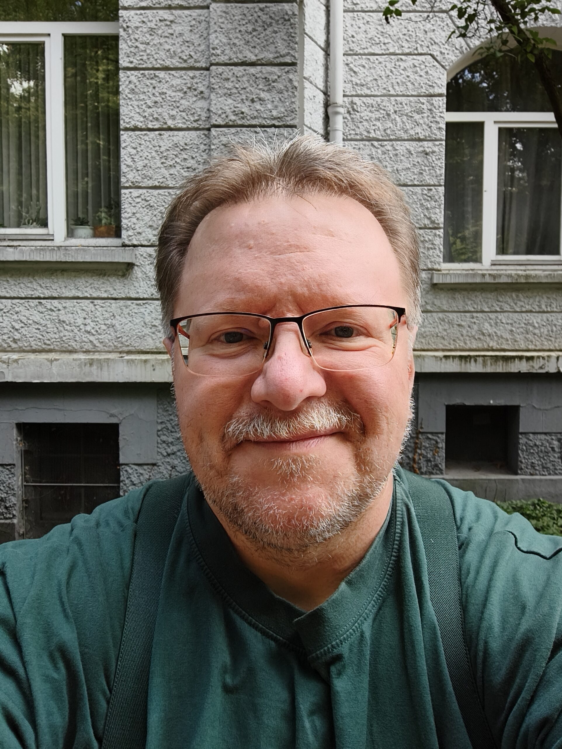 Porträtfoto im Freien: Eine Person mit kurzen Haaren und einer Brille blickt die betrachtende Person direkt an. Die Person hat eine mehrtägige Gesichtbeharrung. Die Person trägt ein waldgrünes T-Shirt und farblich passende Hosenträger. Im Hintergrund eine Häuserwand in Steinoptik und Teile von Fenstern.